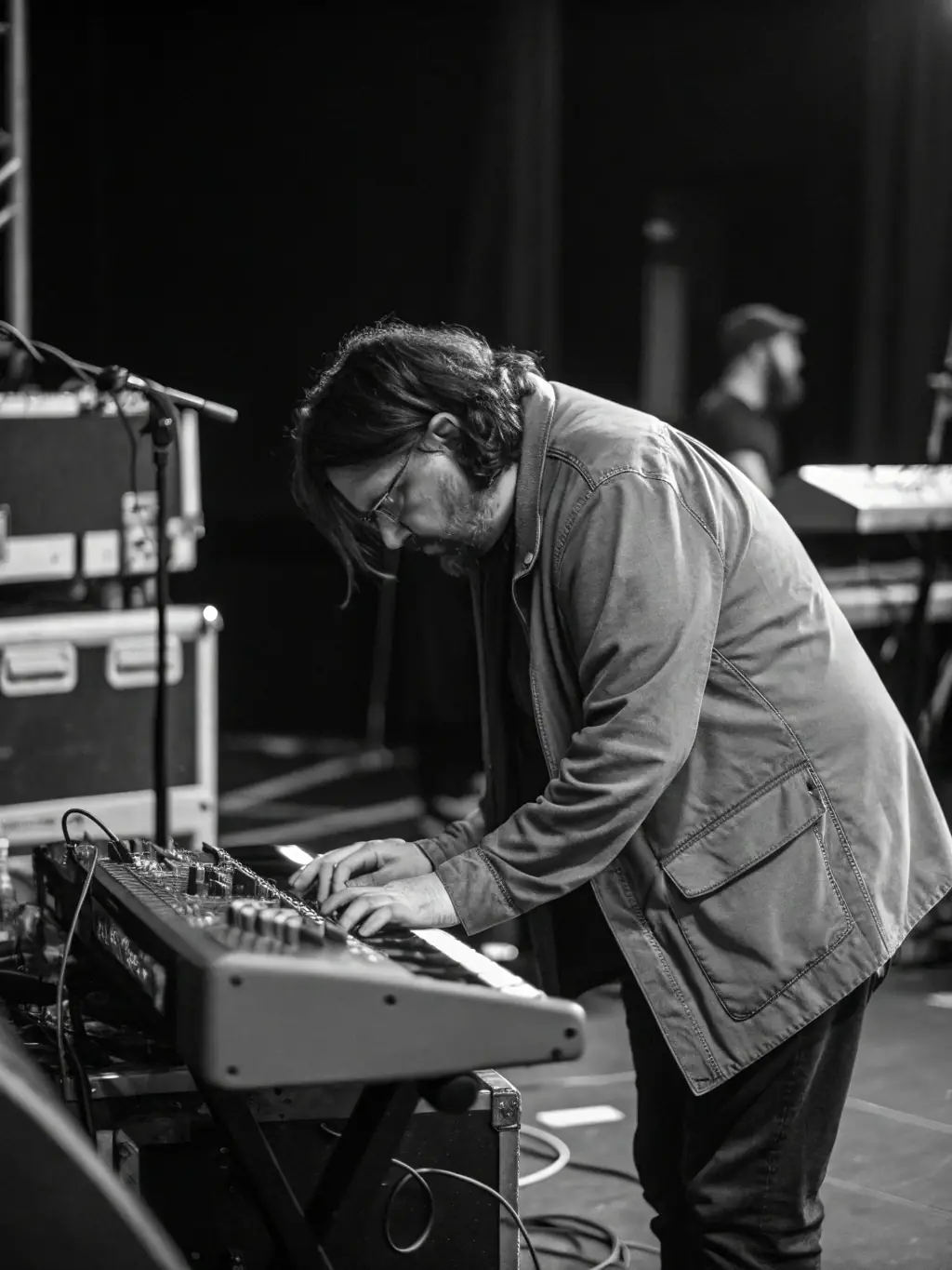 A black and white photo of a KERMHIT ASSO team member providing technical assistance to a band during a soundcheck at a local venue.